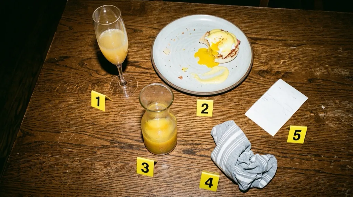 Overhead view of a wooden restaurant table after brunch with a mimosa, a half-eaten eggs benedict, a carafe of orange juice, a striped napkin, and a receipt, each next to a yellow numbered evidence marker