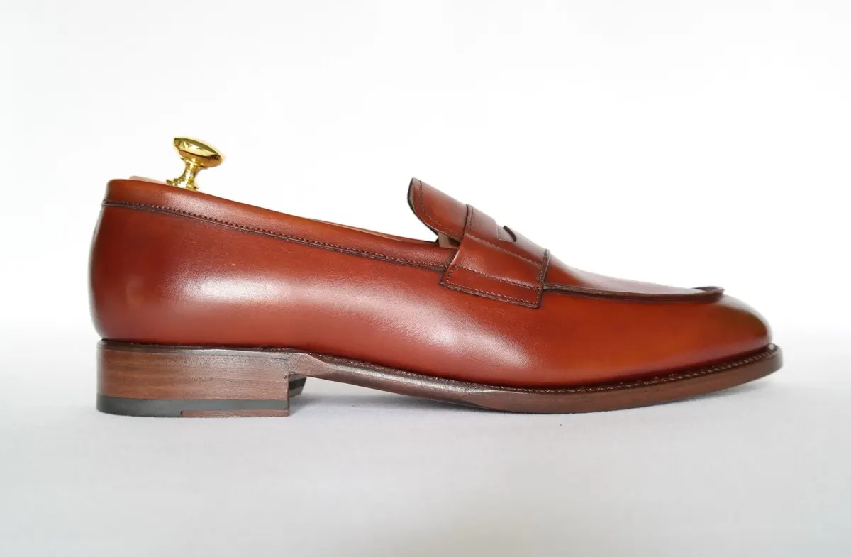 A single brown leather penny loafer with a wooden shoe tree, photographed in profile against a white background