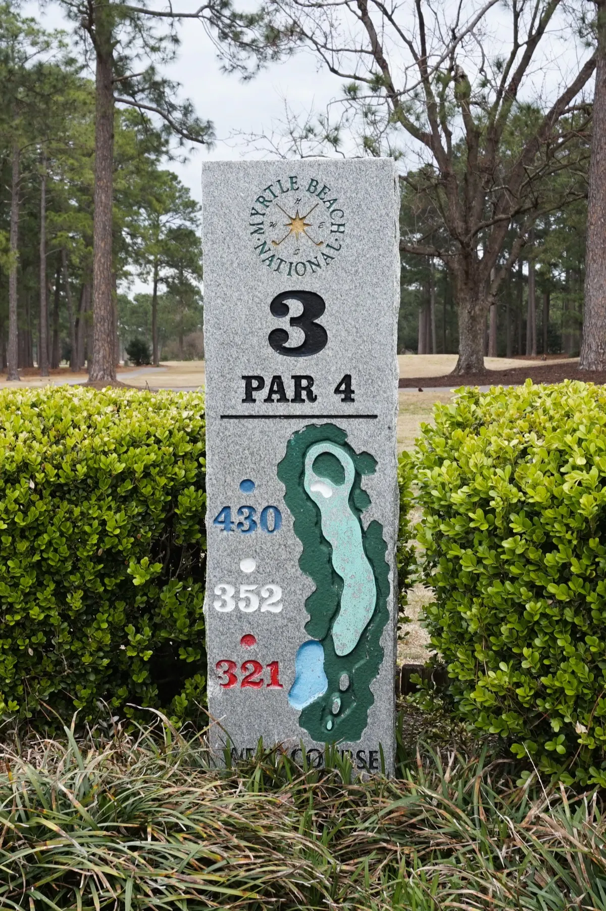 A granite tee marker for hole 3, a par 4, on a Myrtle Beach golf course, set between two boxwood shrubs with pine trees behind it