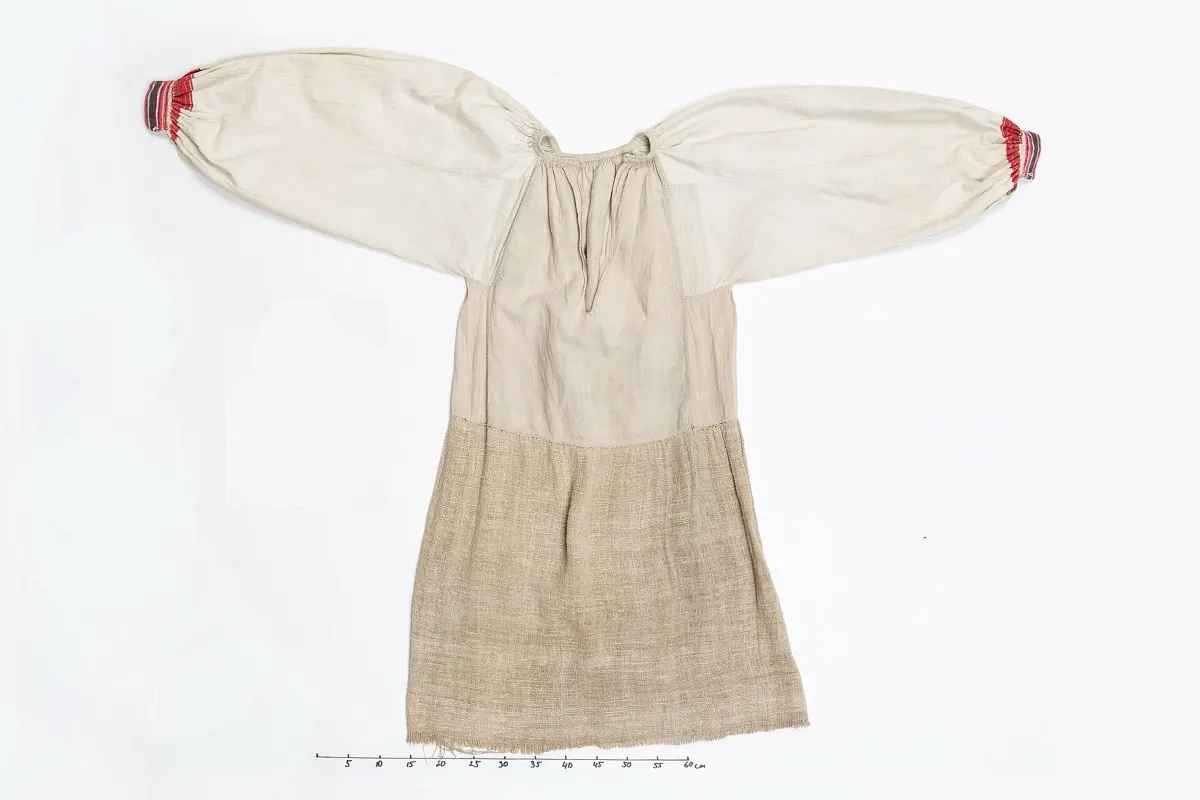 A historical handwoven linen shirt with red embroidered cuffs, photographed flat on a white background with a measuring scale