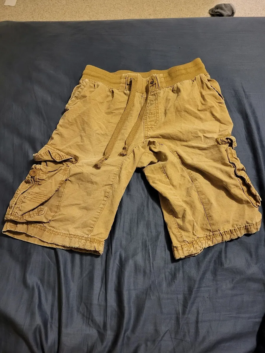 A pair of faded khaki cargo shorts with a drawstring waist and large side pockets, laid flat on a blue bedsheet