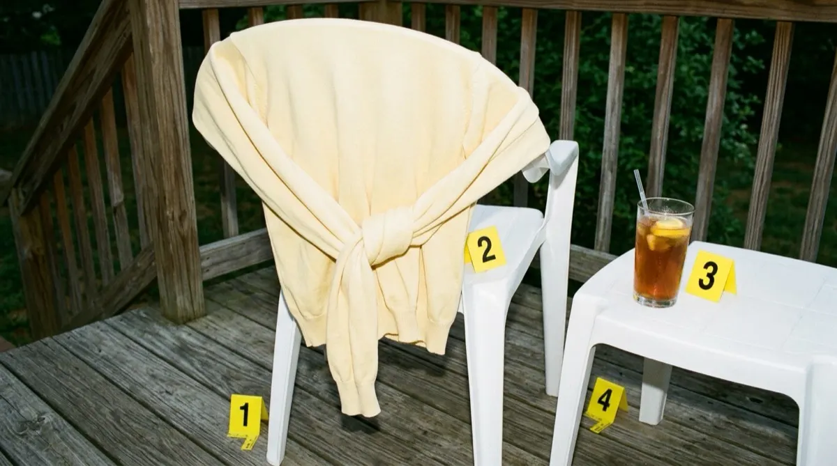 A pale yellow cotton sweater knotted by its sleeves around the back of a white plastic patio chair on a wooden deck, next to a glass of iced tea, with yellow numbered evidence markers